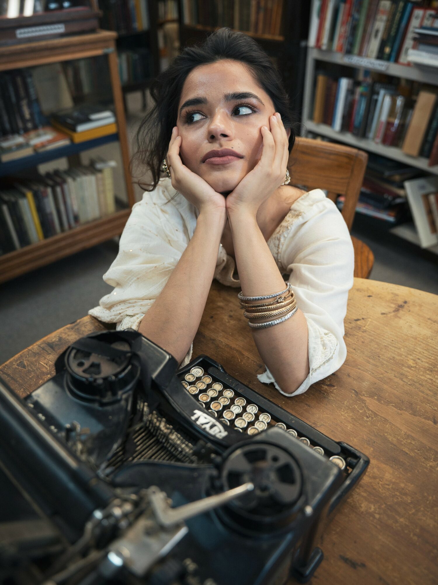 Woman with dark hair resting her face on her hands, sitting at a table with a vintage typewriter and a camera, surrounded by books.