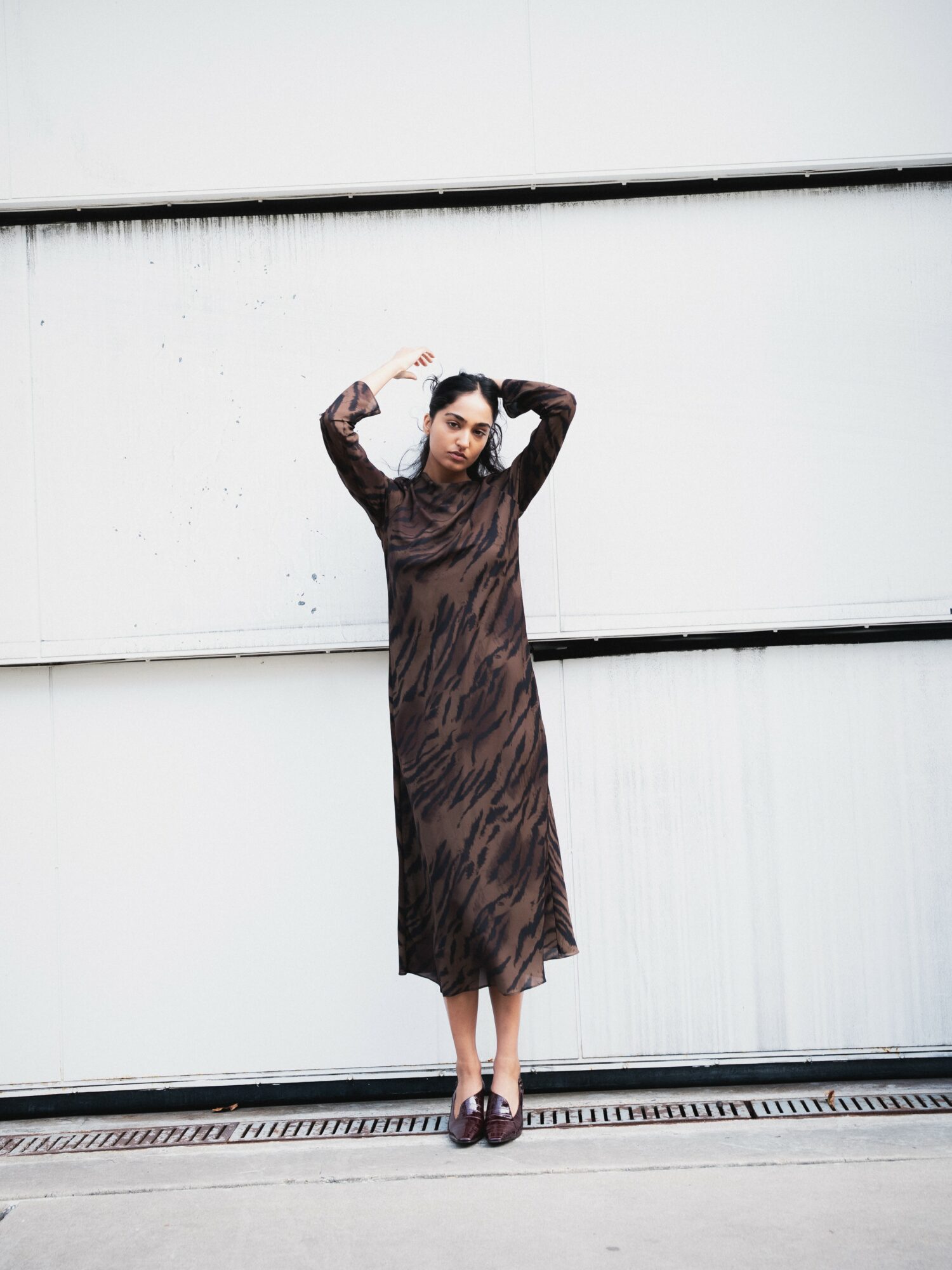 Woman in a long, dark patterned dress standing against a white wall with her hands raised above her head.