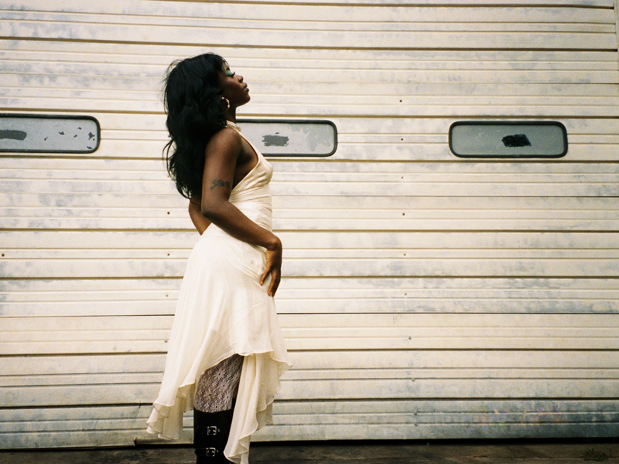 Woman in a cream dress standing sideways against a metal garage door with small windows.