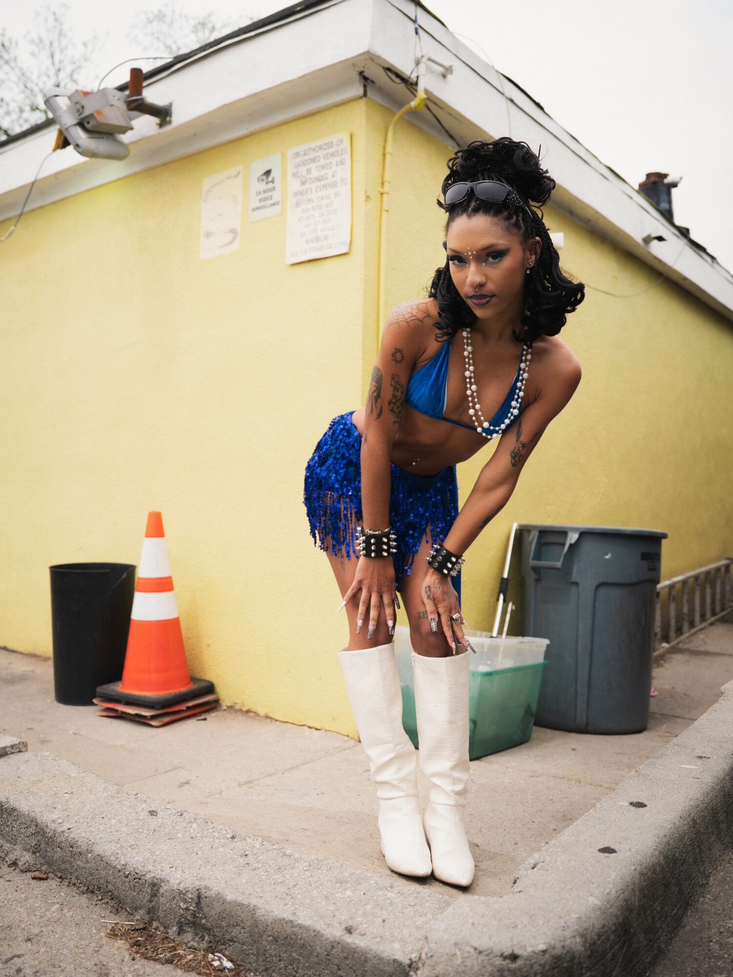 Woman with dark curly hair in a blue outfit and white boots standing on sidewalk near yellow building.