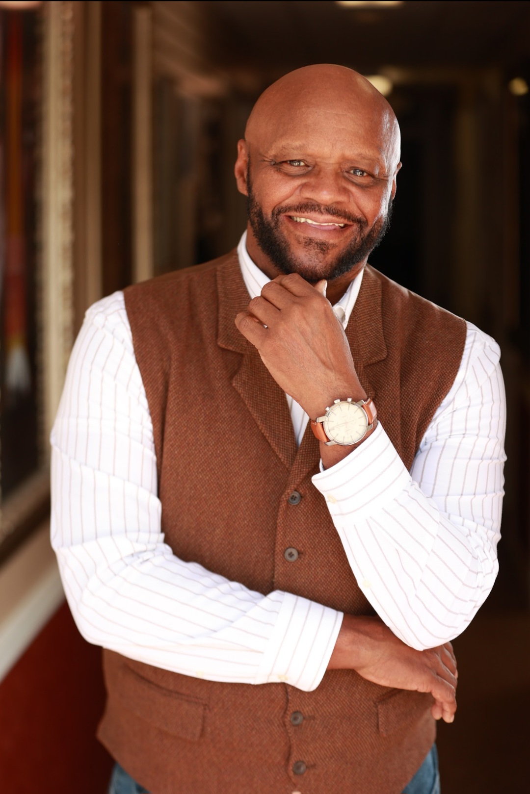 Man with a beard smiling, wearing a brown vest over a white shirt, standing indoors.