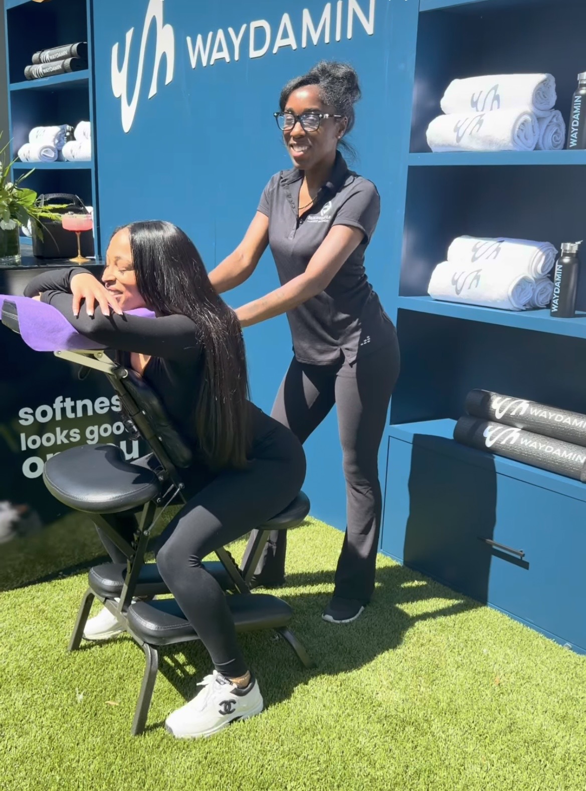 Two women at a massage station, one standing and smiling, the other seated with eyes closed, in front of a blue display with towels.