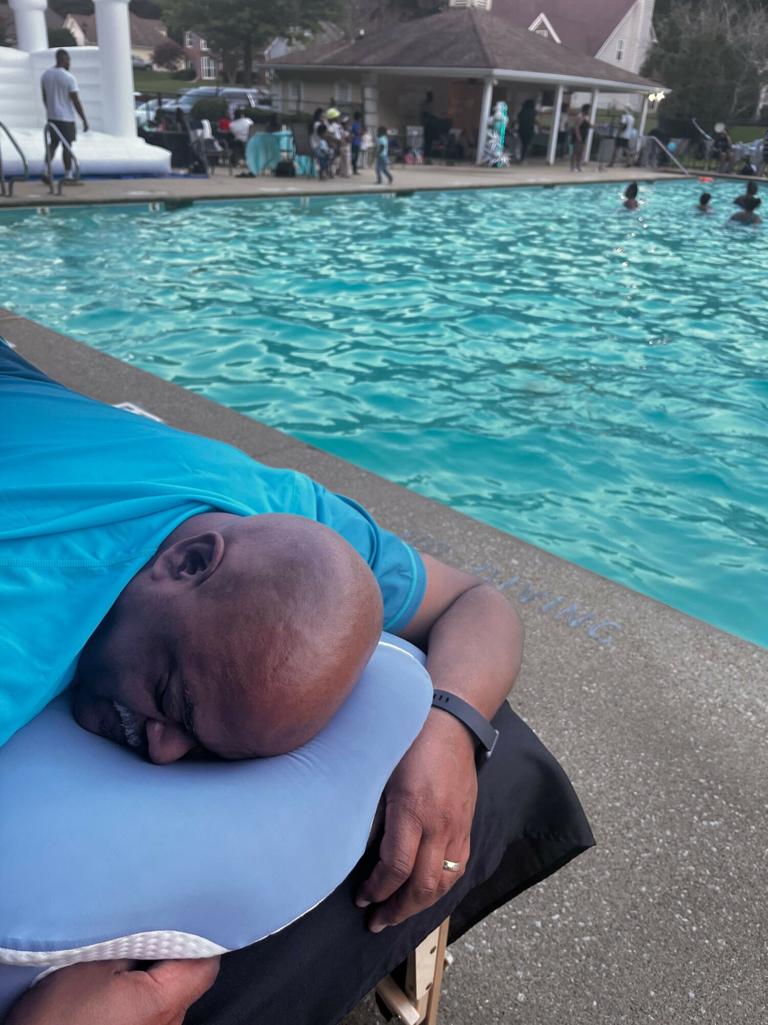 Person resting by poolside with head on a pillow, wearing a blue shirt, near swimming pool with people in background.