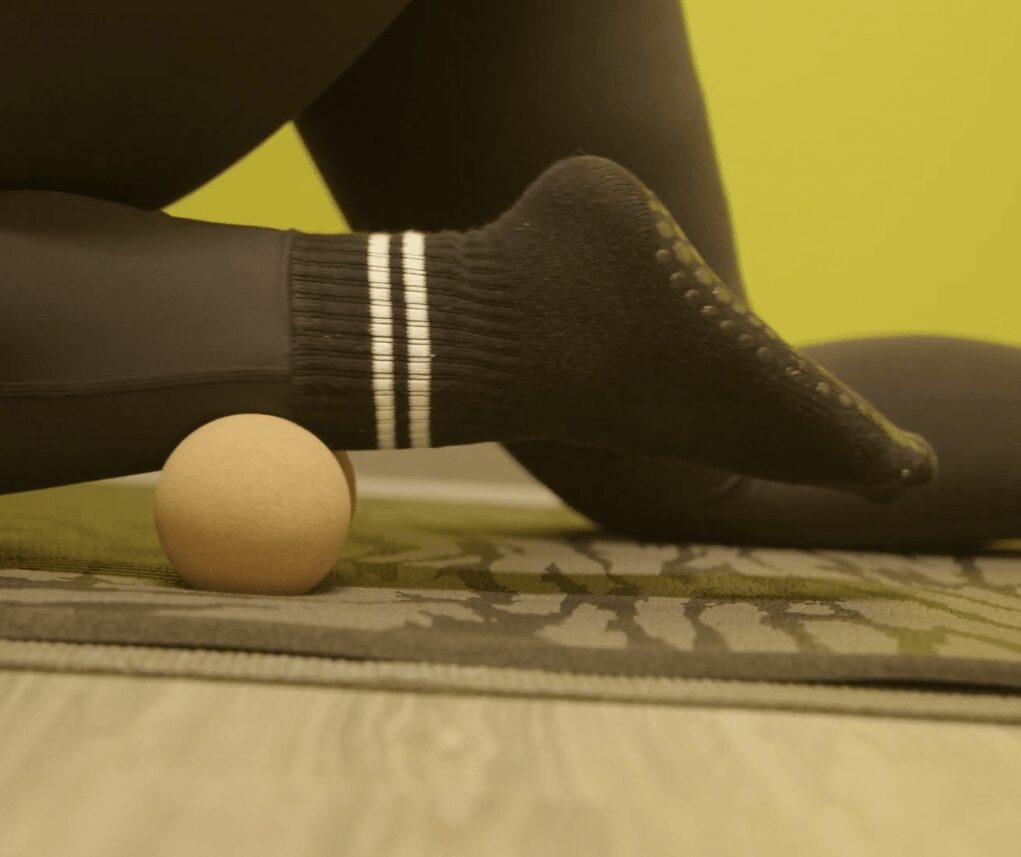 Person stretching foot on a yoga mat with a ball nearby, against a yellow wall.