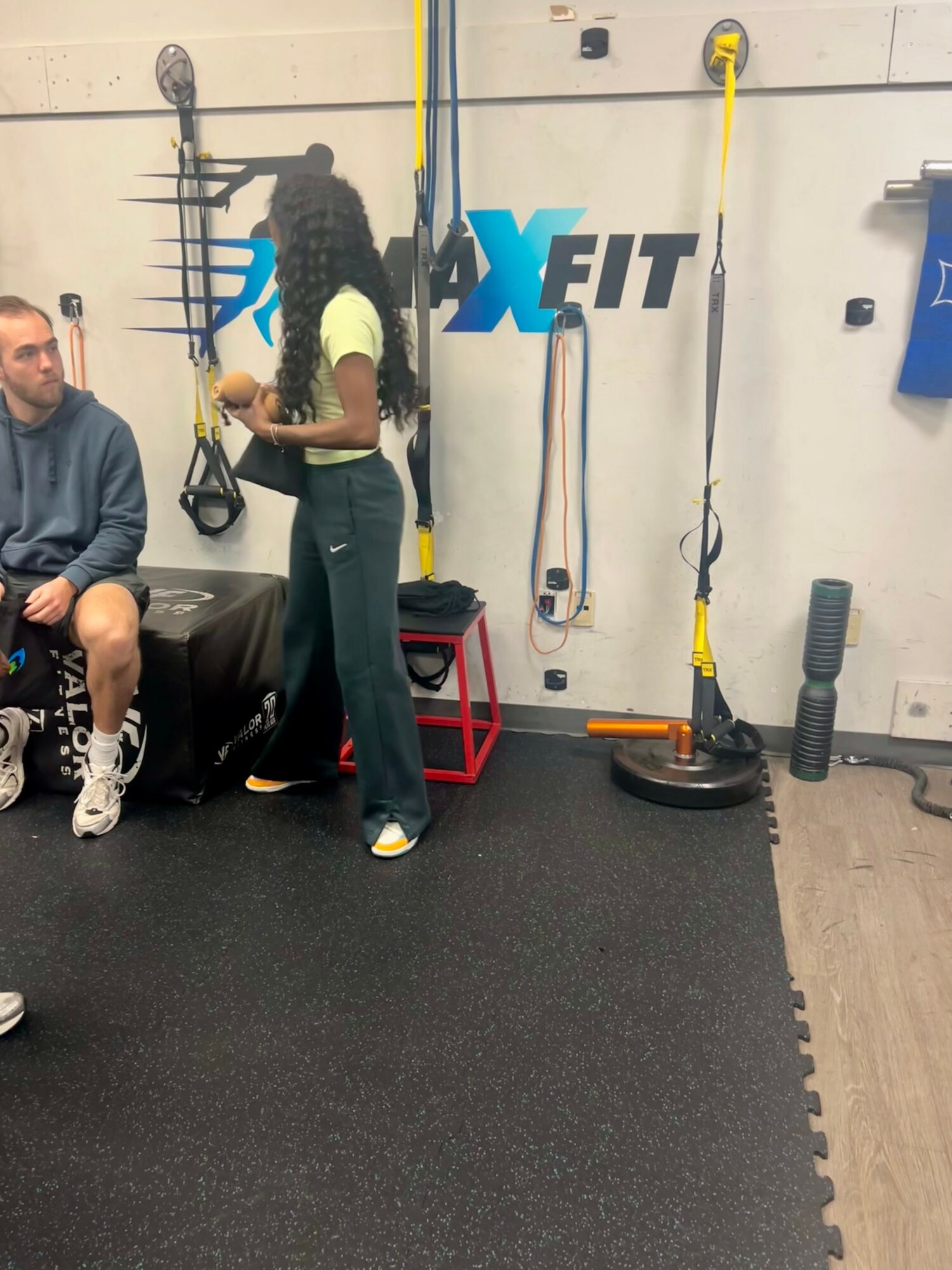Person standing in gym, others sitting on bench, gym equipment and wall with logo in background.