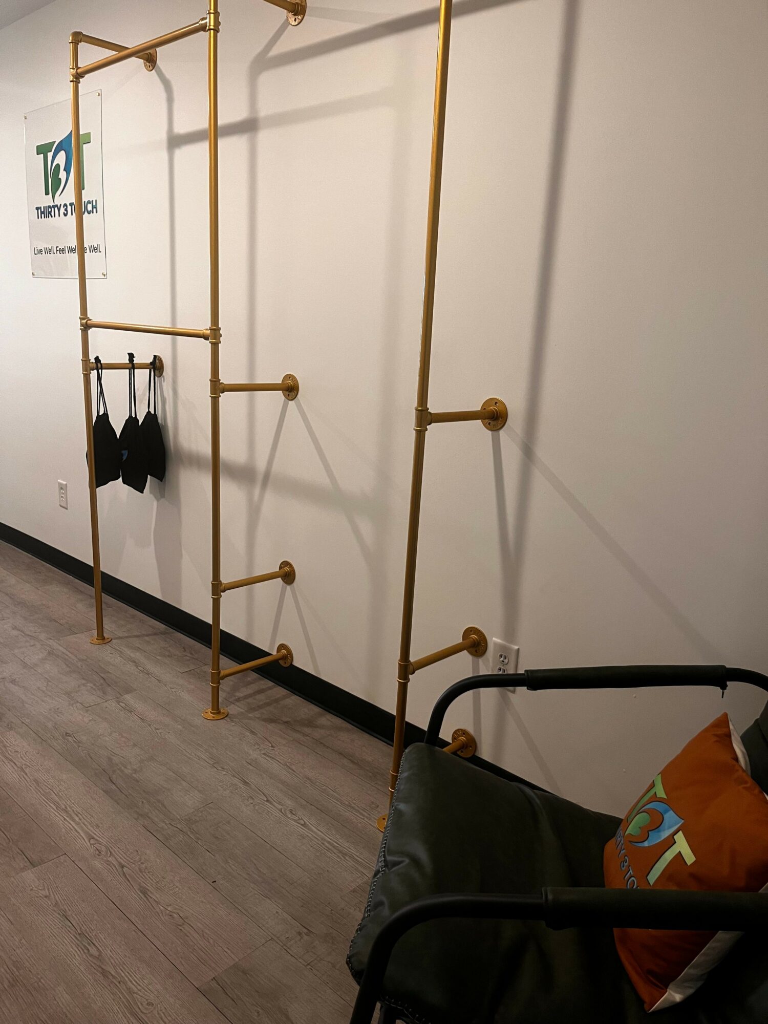 Gold-colored wall-mounted clothing rack with hooks and bars, with black bags hanging, in a room with white walls and wooden floor.