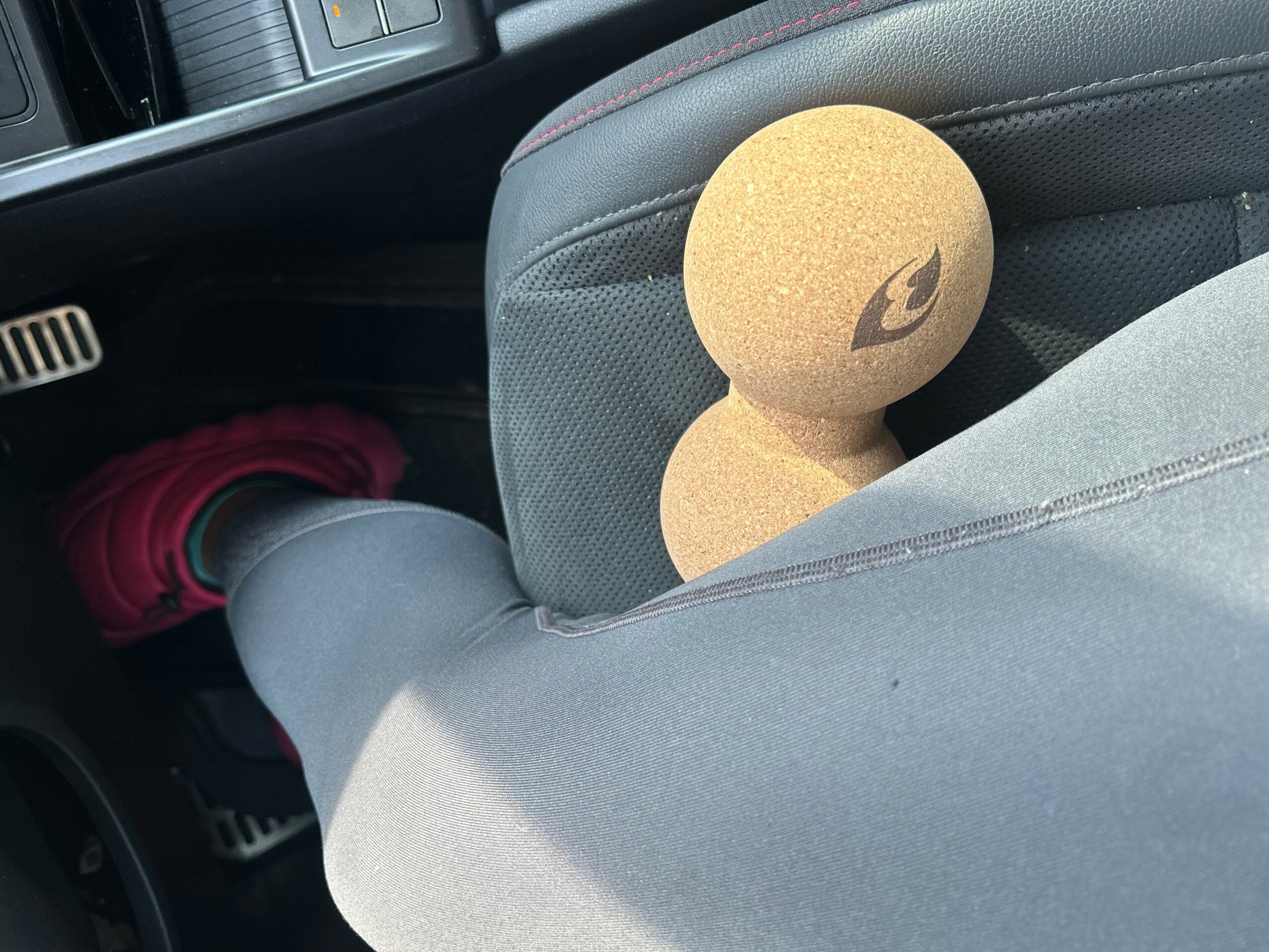 A plush toy with a leaf logo rests on a car seat next to a person's leg wearing gray pants and red shoes.