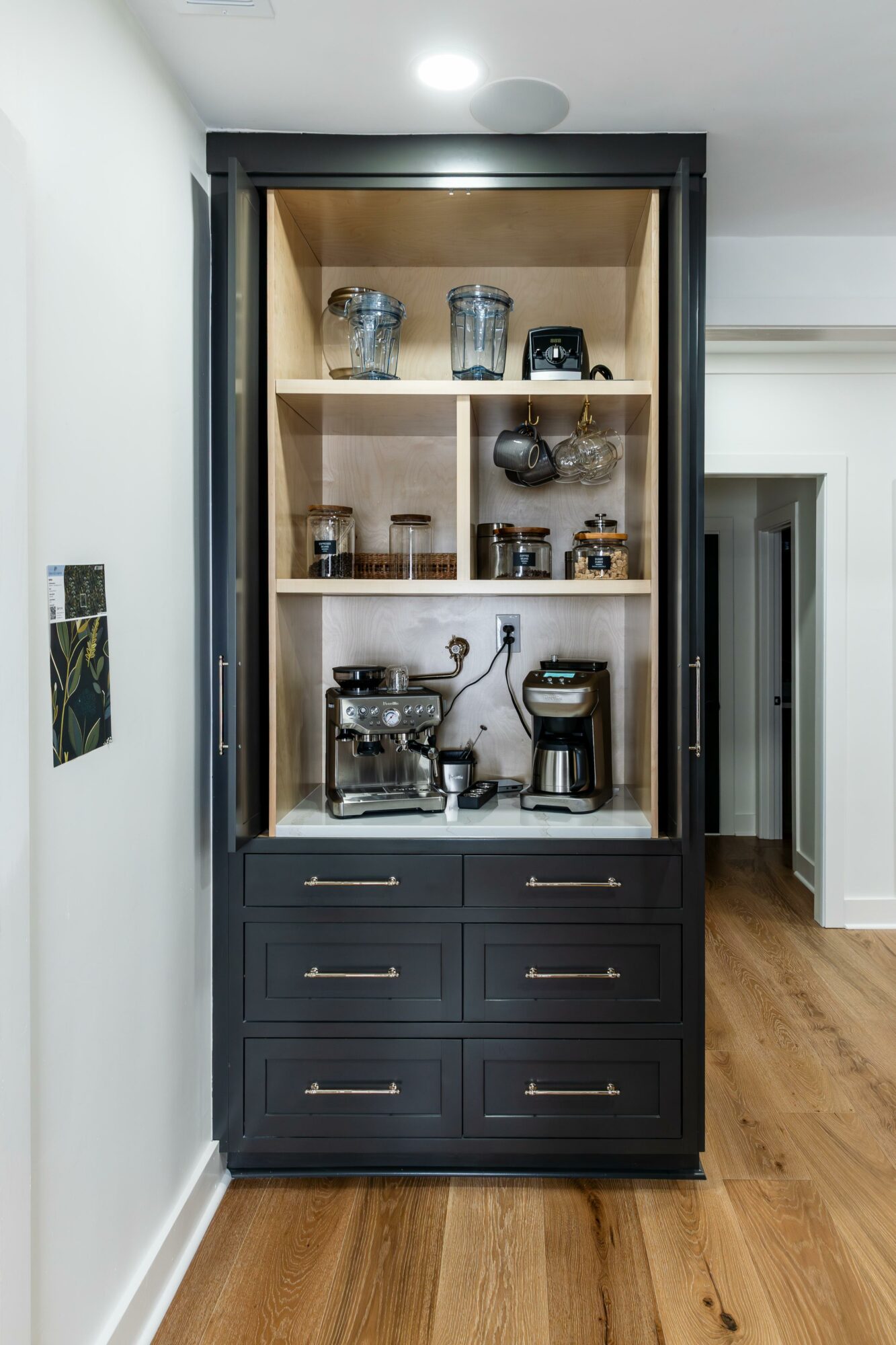 Tall black cabinet with glass doors, wooden shelves, coffee machine, and jars inside, in a room with white walls and wooden floor.