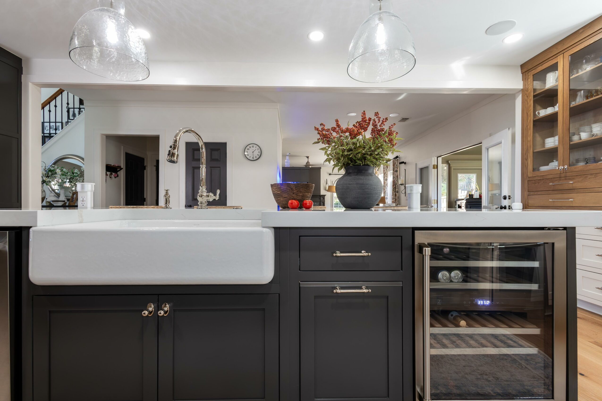 Modern kitchen with black cabinets, white countertop, sink, flowers, and glass-front cabinet, open living space in background.