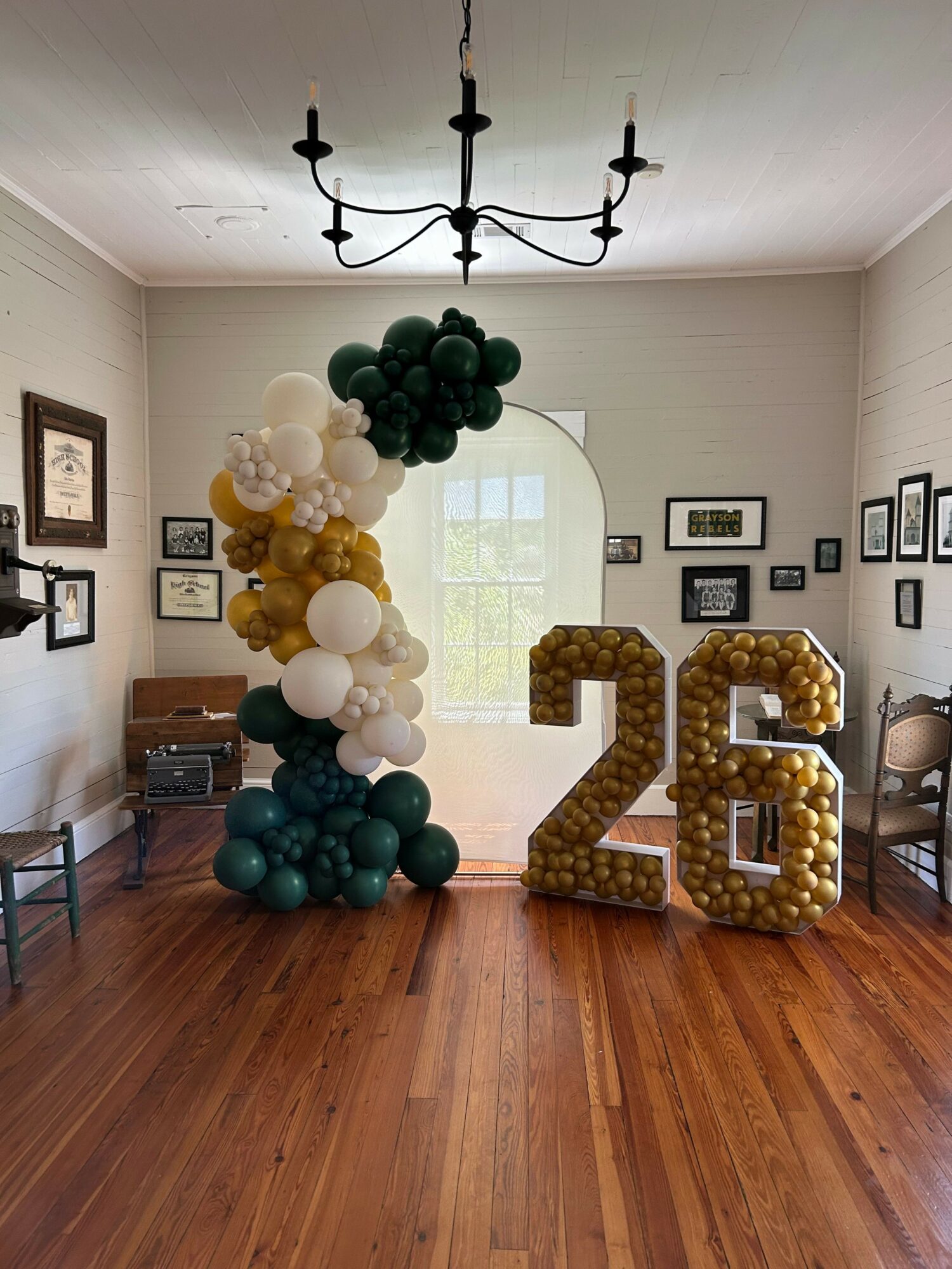Balloon arch, large number 26, and a window in a decorated room with framed pictures on walls.