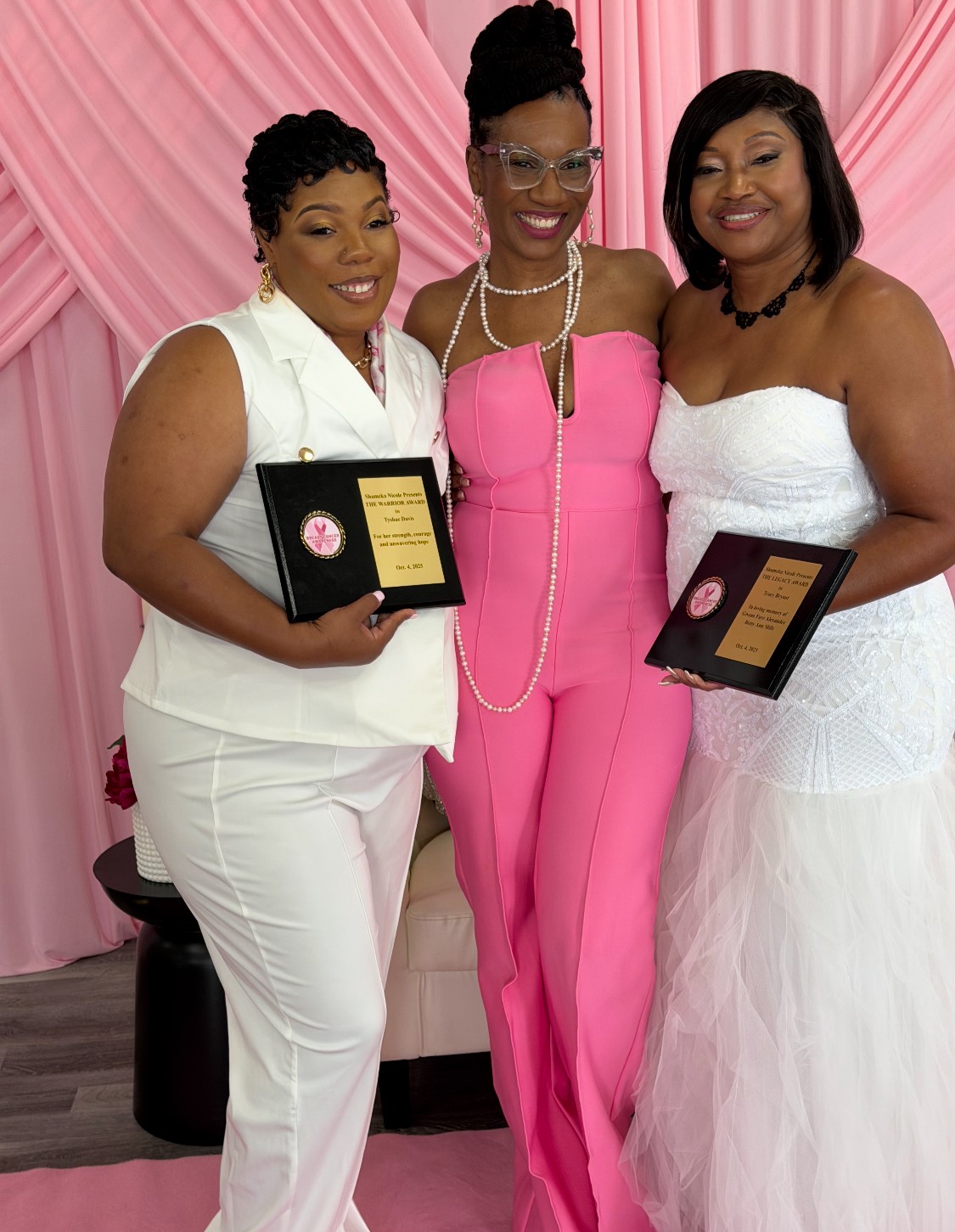 Three women in elegant dresses holding awards, standing in front of pink draped background.