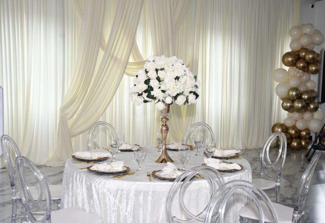 Round table with white tablecloth, floral centerpiece, and transparent chairs in a decorated room with draped curtains and balloons.