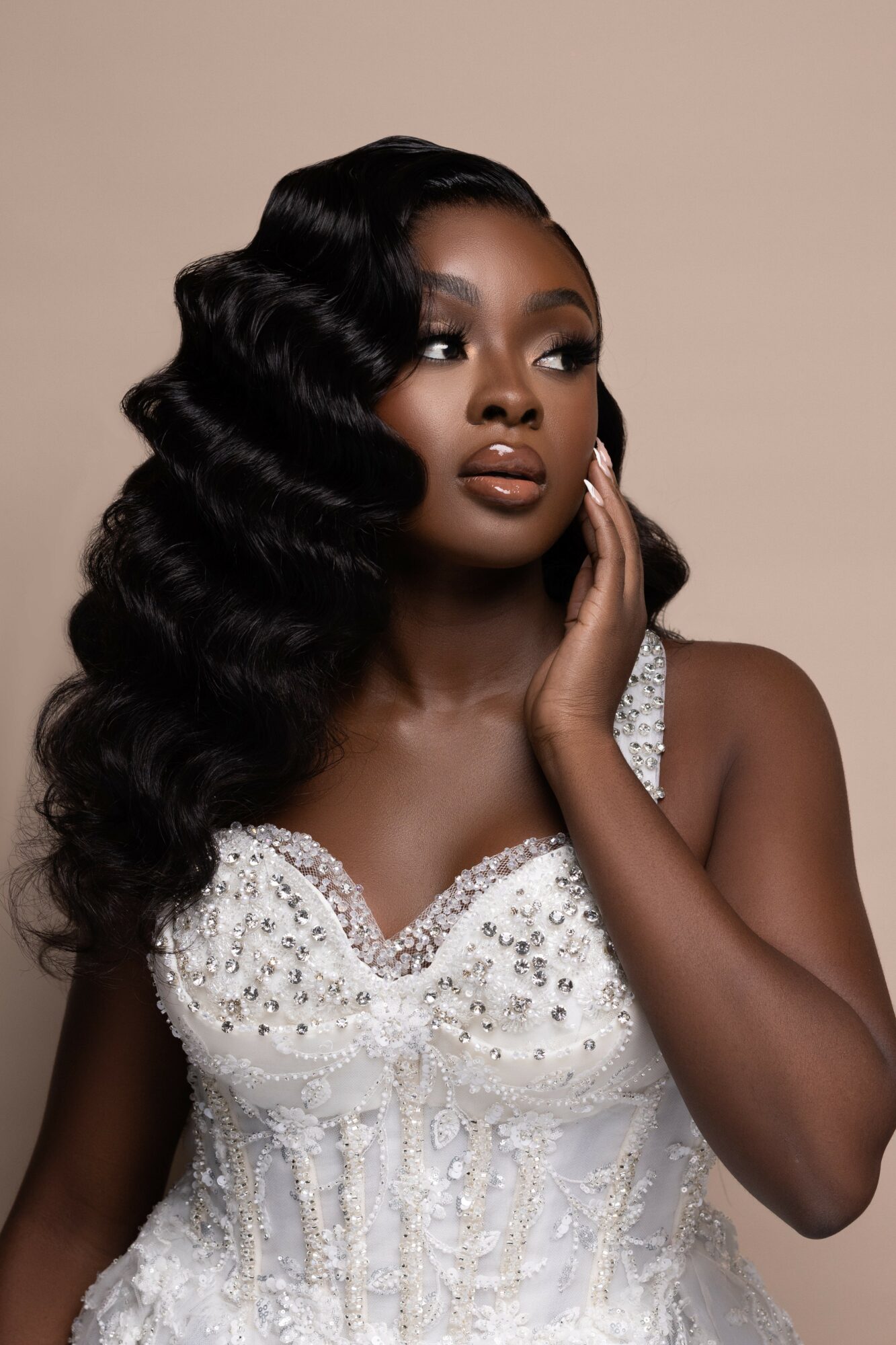 Woman with long, wavy black hair wearing a white, beaded dress, touching her face gently, against a plain background.