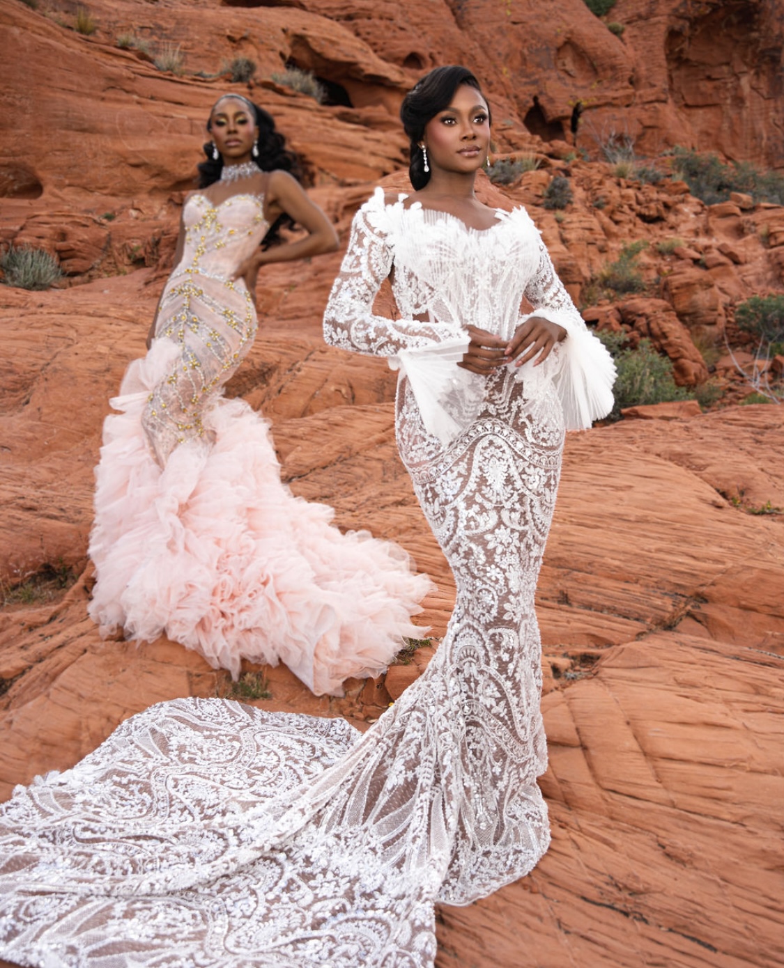 Two women in elegant dresses standing on red rocky terrain with cliffs in background.