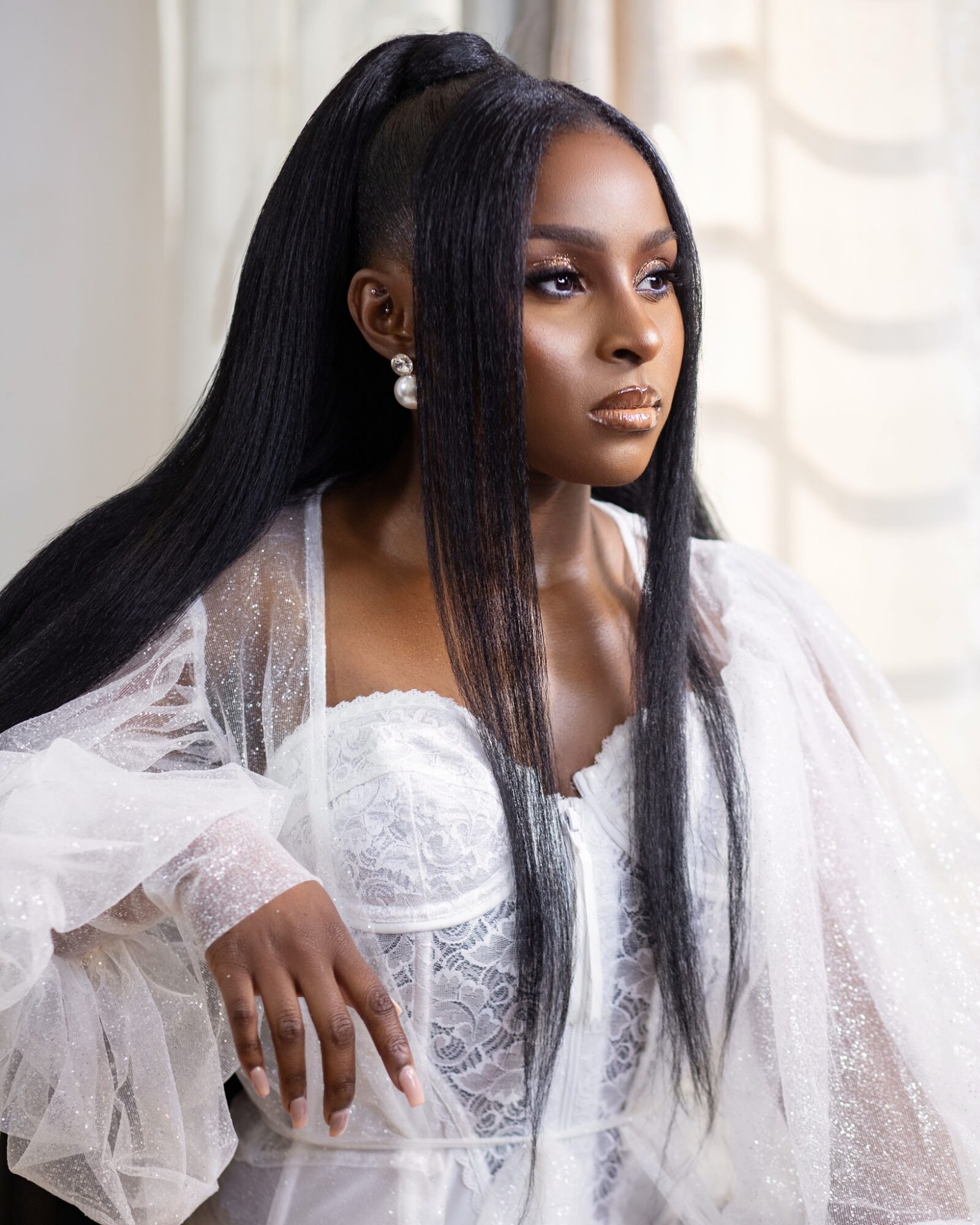 Woman with long black hair, wearing a white lace dress and pearl earrings, looking to the side.