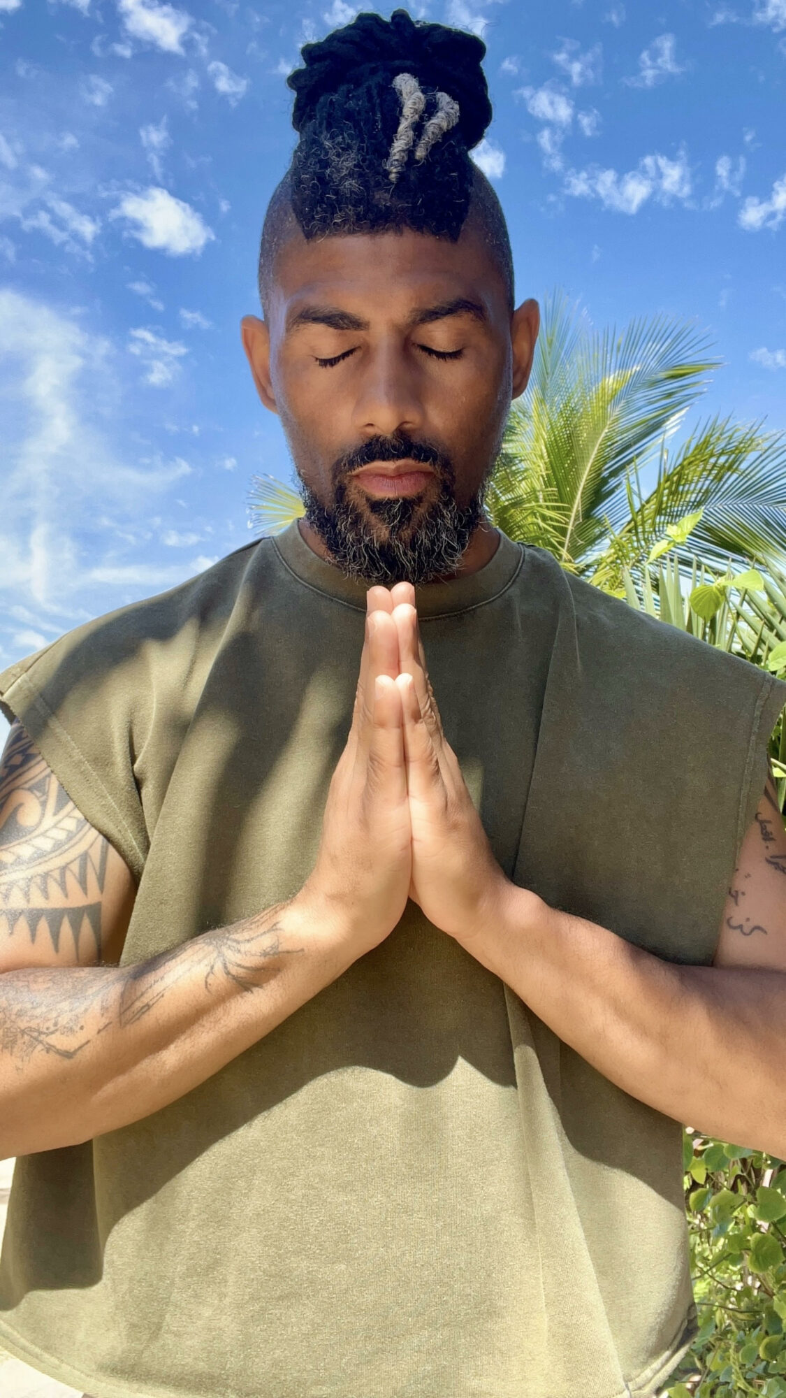 Man with closed eyes and hands pressed together in prayer outdoors, blue sky and palm trees in background.