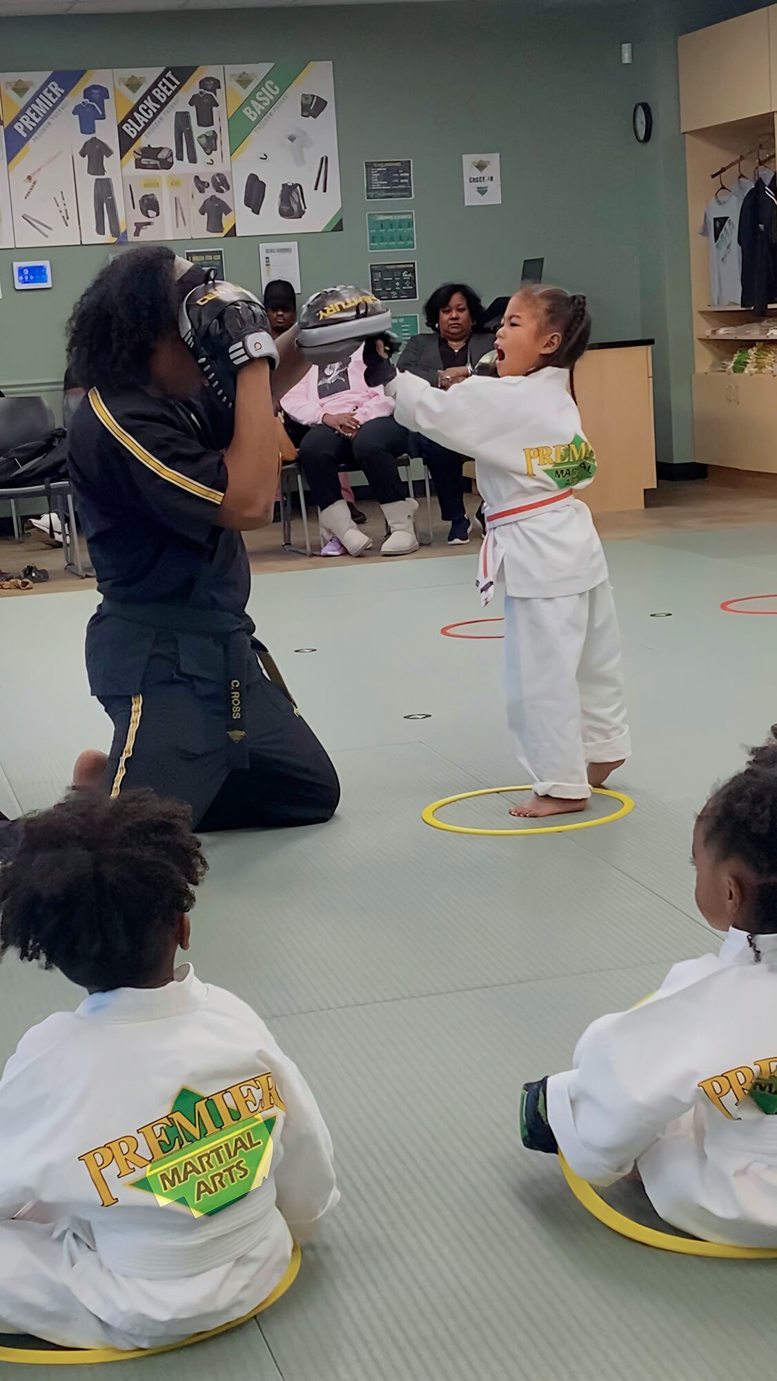 Child in a martial arts uniform receiving a certificate from an instructor in a dojo with children sitting on the floor.