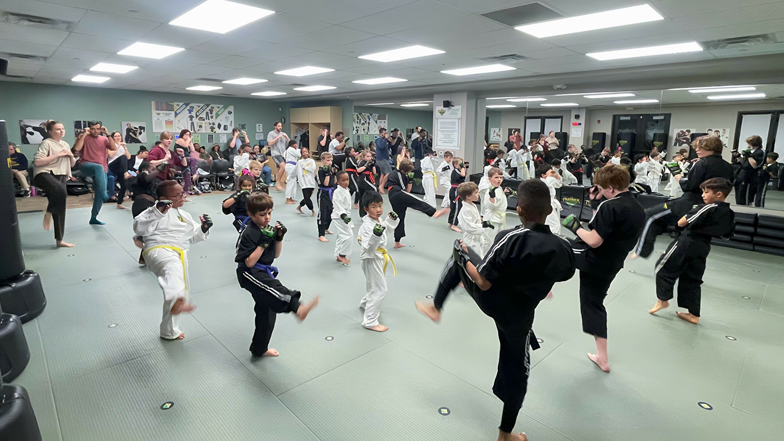 Group of children practicing martial arts in a spacious indoor gym with adults observing.