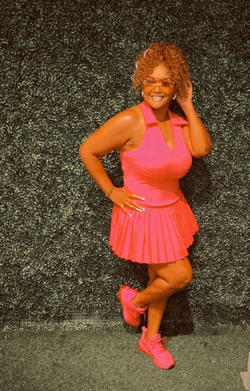 Woman with curly hair in pink dress and sneakers, smiling, standing against a dark textured background.