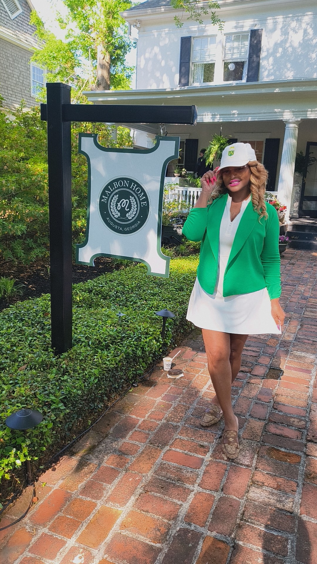 Woman in green blazer, white dress, and white hat walking on brick path outside building with sign and greenery.