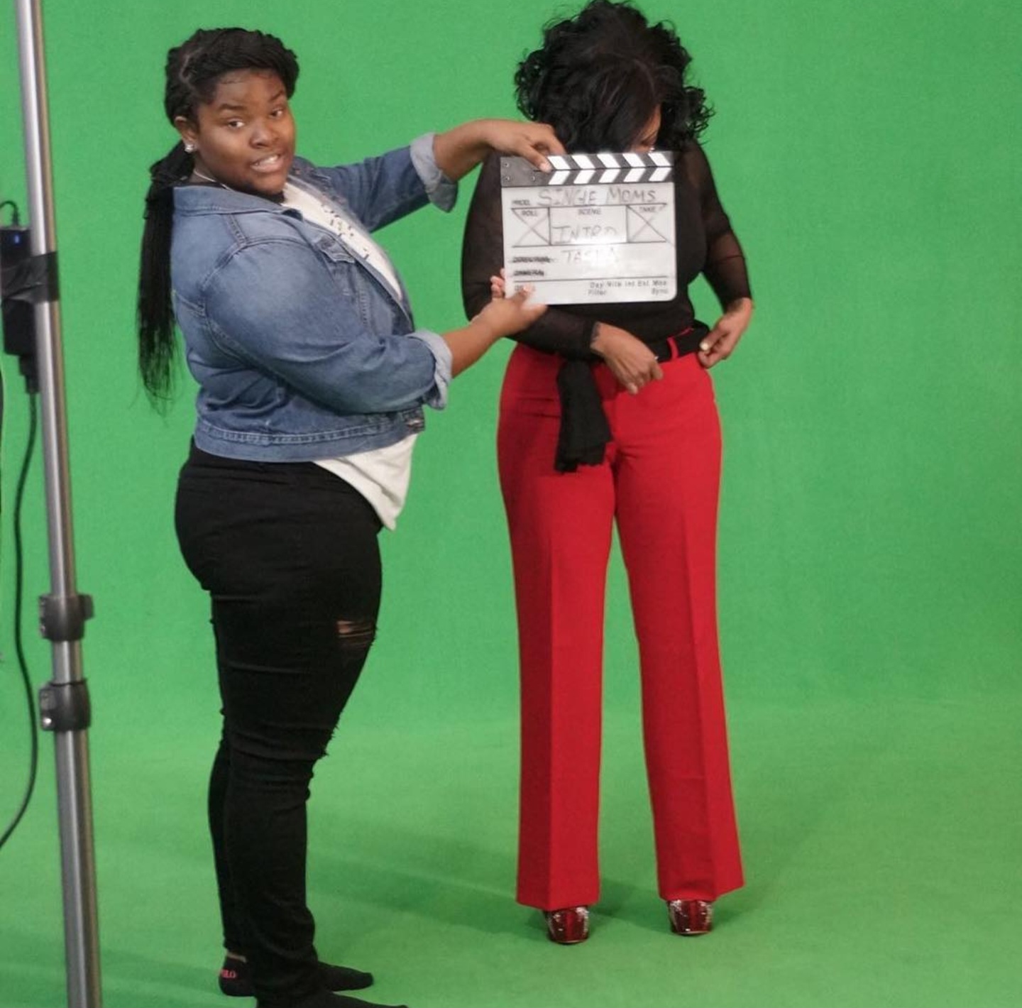 Two women standing in front of a green screen, one holding a film slate, the other with hands on hips.