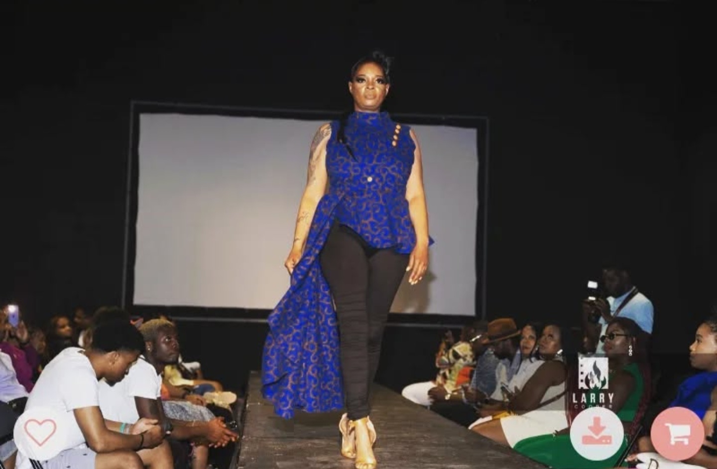 Model walking on runway with audience seated on both sides, large screen behind, wearing blue and black outfit.