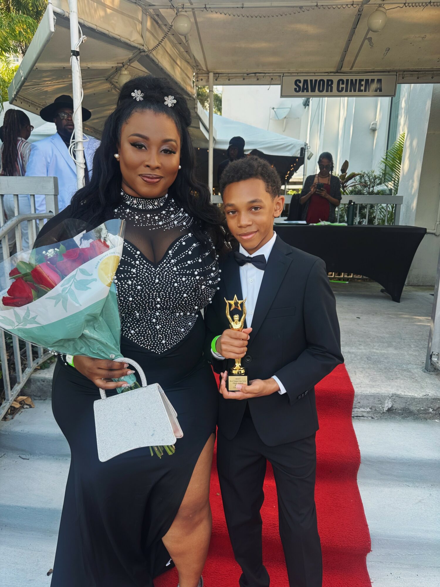 Woman in black dress holding a bouquet of flowers and a white purse, standing next to a young boy in a tuxedo holding a trophy.