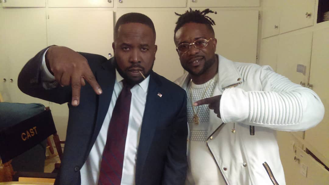 Two men pose indoors, one in a suit and the other in a white jacket, both pointing downward with their hands.
