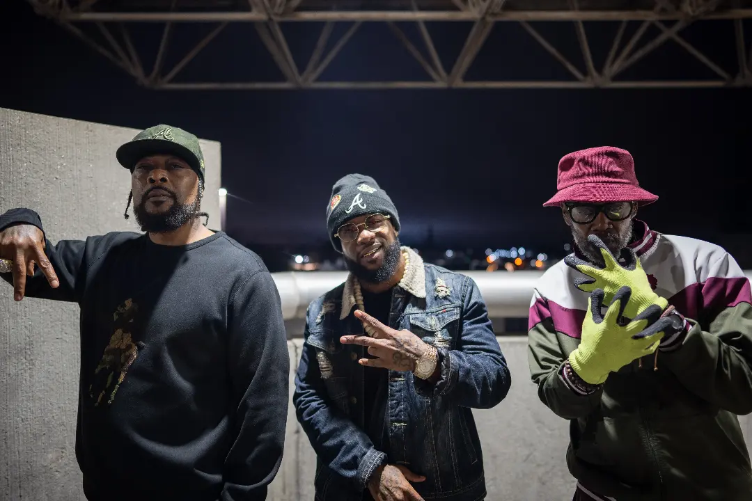 Three men standing outdoors at night, one making a peace sign, another wearing a red hat and yellow gloves, city lights in background.