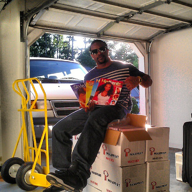 Man sitting on boxes inside a garage holding magazines, with a yellow hand truck nearby, sunlight outside.