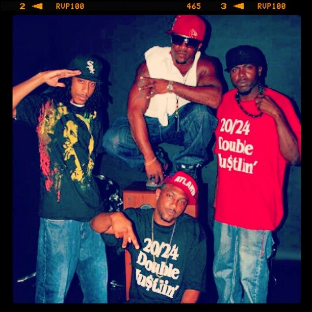Four men pose together, two wearing caps with one making a salute gesture, two wearing shirts with text, in a dark setting.