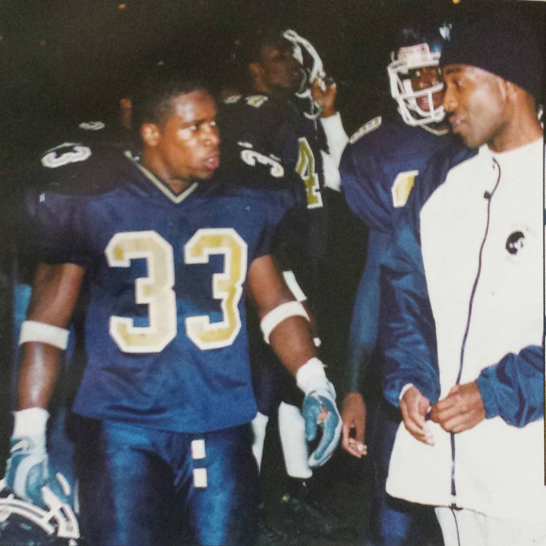 Group of football players and a coach in dark uniforms, some wearing helmets, standing outdoors at night.