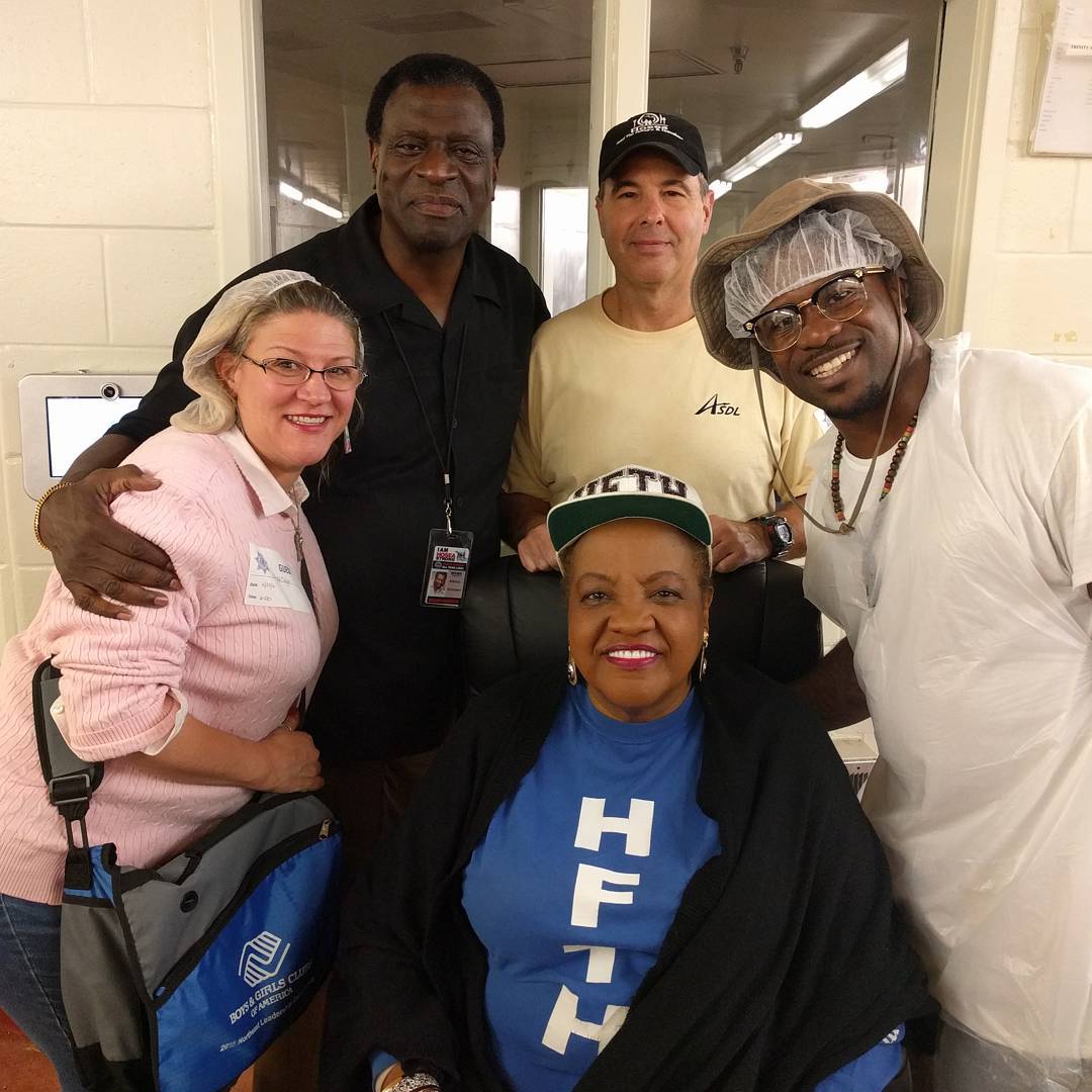 Group of five people smiling indoors, some wearing hats and glasses, standing close together.