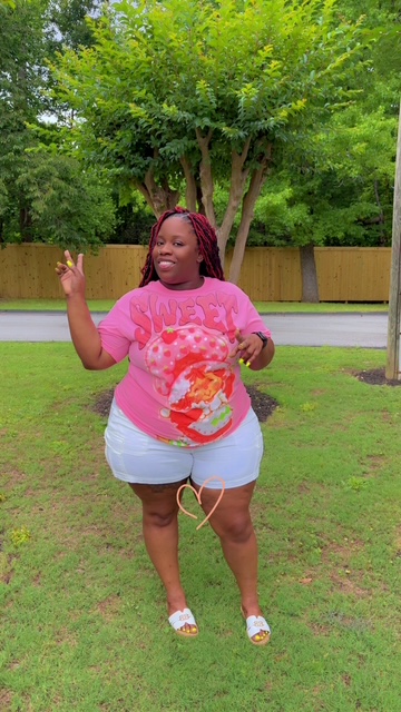 Person standing outdoors on grass, wearing a pink T-shirt and light blue shorts, with trees and a wooden fence in background.