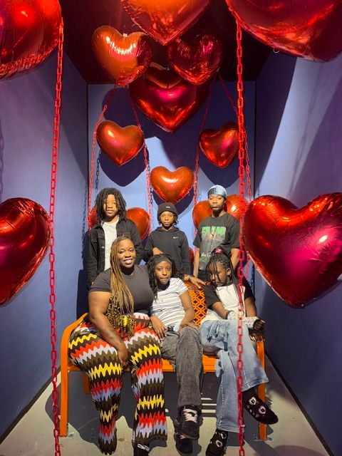 Group of people standing in front of red heart-shaped balloons, some holding balloons, in a decorated room.
