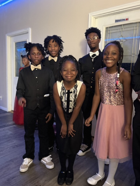 Group of five children dressed in formal attire, standing indoors in front of a window and door, smiling.