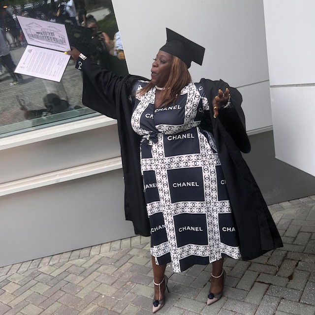 Woman in graduation gown and cap holding diploma outside building, smiling, with glass window and white wall behind her.
