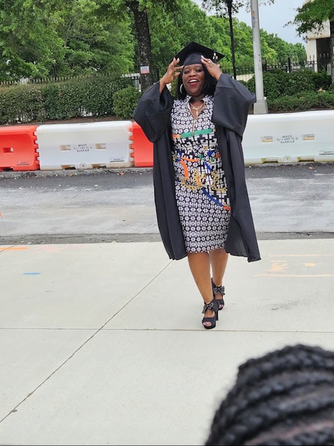 Person in graduation gown and cap smiling outdoors, holding cap with both hands, standing on sidewalk with trees and barriers behind.
