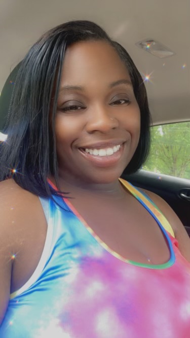 Smiling woman with shoulder-length black hair in a colorful tie-dye top inside a car.