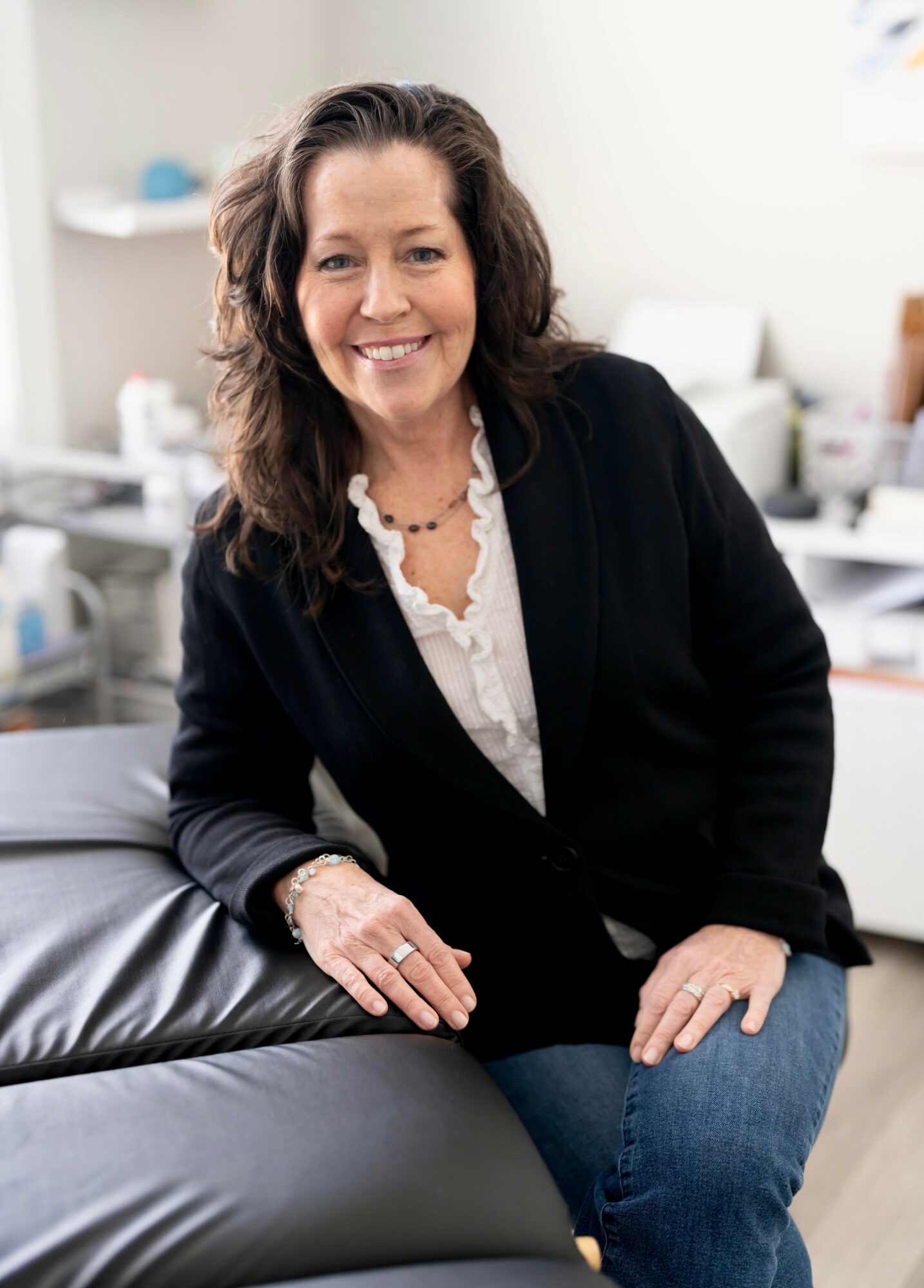 Woman with dark hair smiling, sitting on a black leather chair in an office, wearing a black blazer and white blouse.