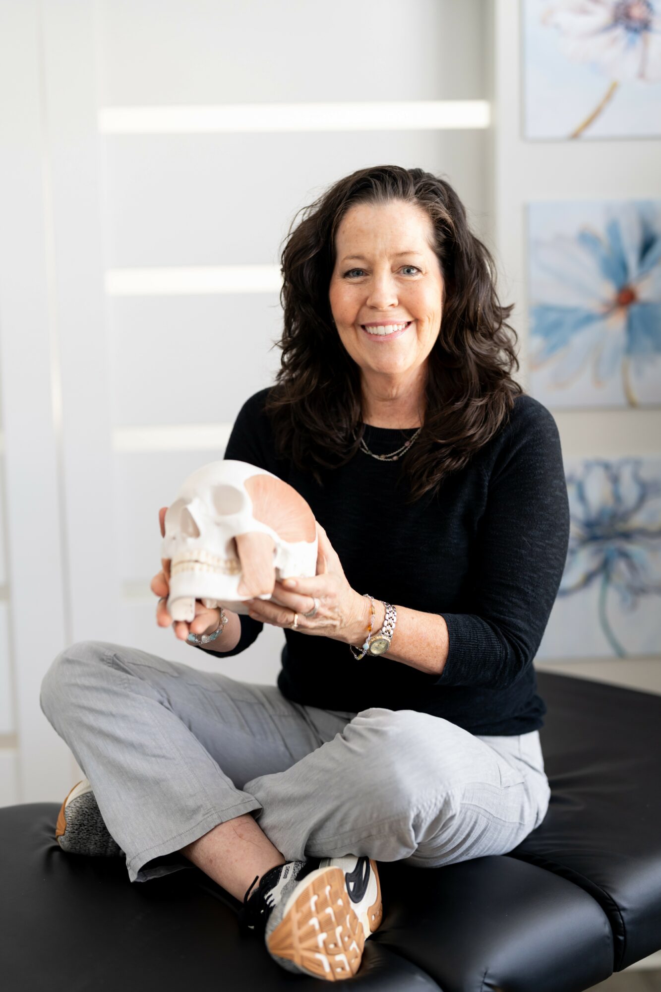 Woman sitting cross-legged on a black bench, holding a skull model, smiling, in a bright room with artwork on the wall.
