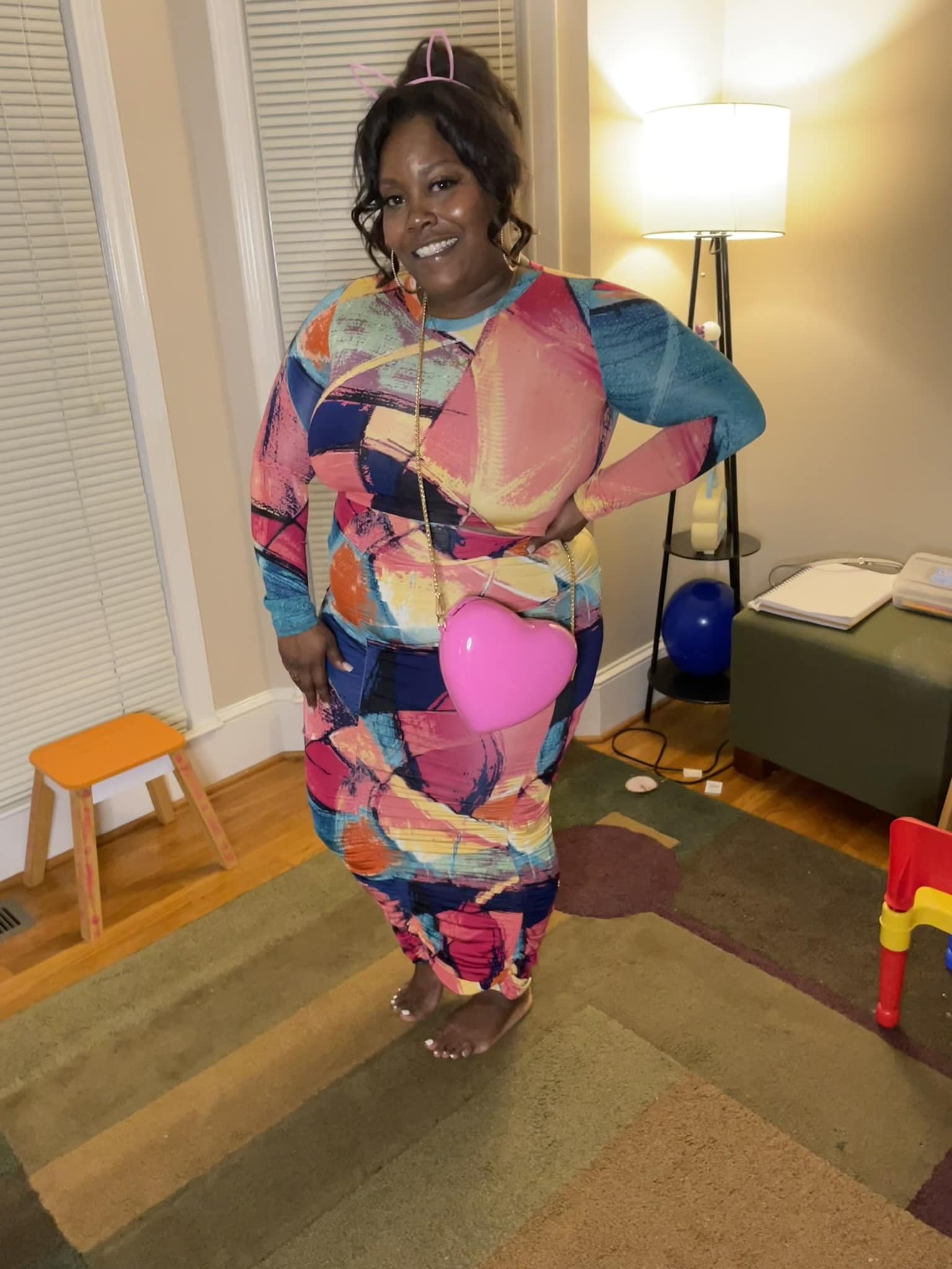 Woman in colorful abstract-patterned outfit holding a pink balloon, smiling indoors with a lamp and furniture in background.