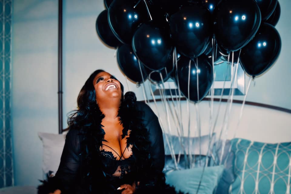 Woman smiling and looking up at black balloons indoors, with pillows and a bed in the background.