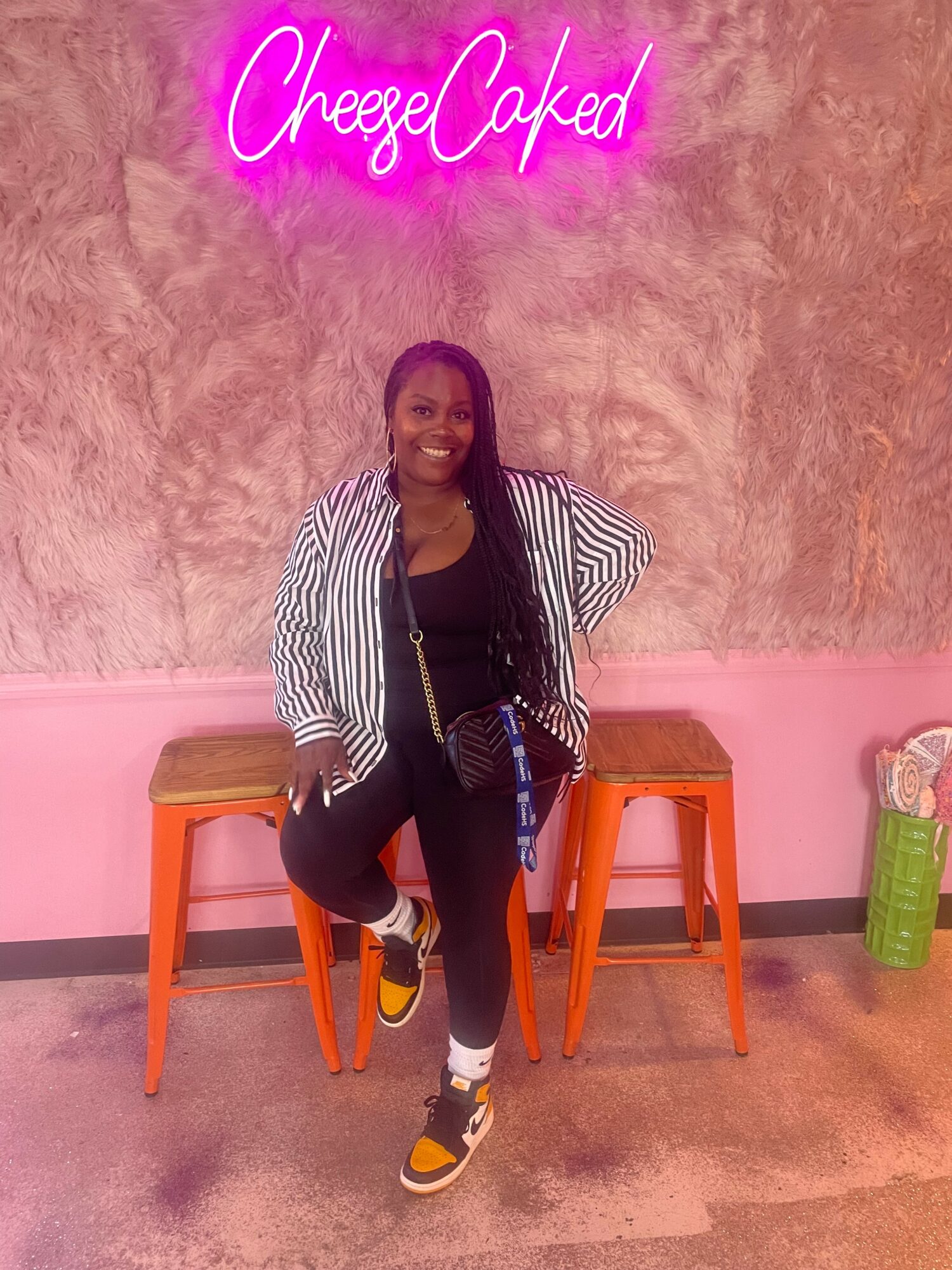 Woman with long hair smiling, standing between two orange stools, pink wall background, neon sign reading 'CheezCaked'.