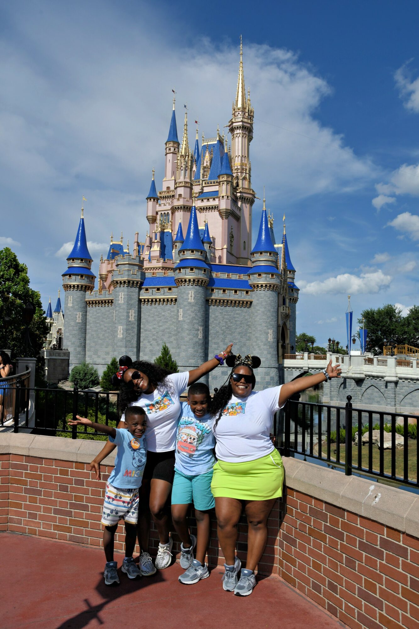 Group of five people posing in front of a castle with blue spires and a brick wall, under a partly cloudy sky.