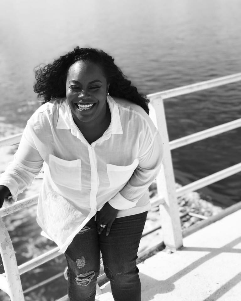 Woman smiling and walking outdoors near water with railing, wearing a white shirt and ripped jeans.