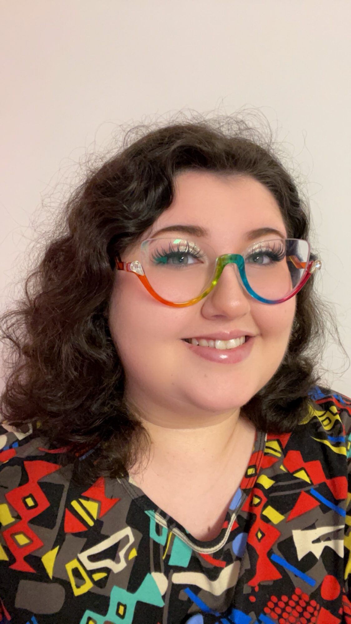 Young woman with curly hair wearing colorful glasses and a patterned shirt, smiling.