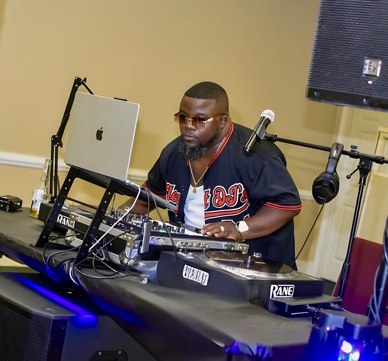 Person wearing glasses and a black sports jersey with red and white accents, working with DJ equipment at a table.