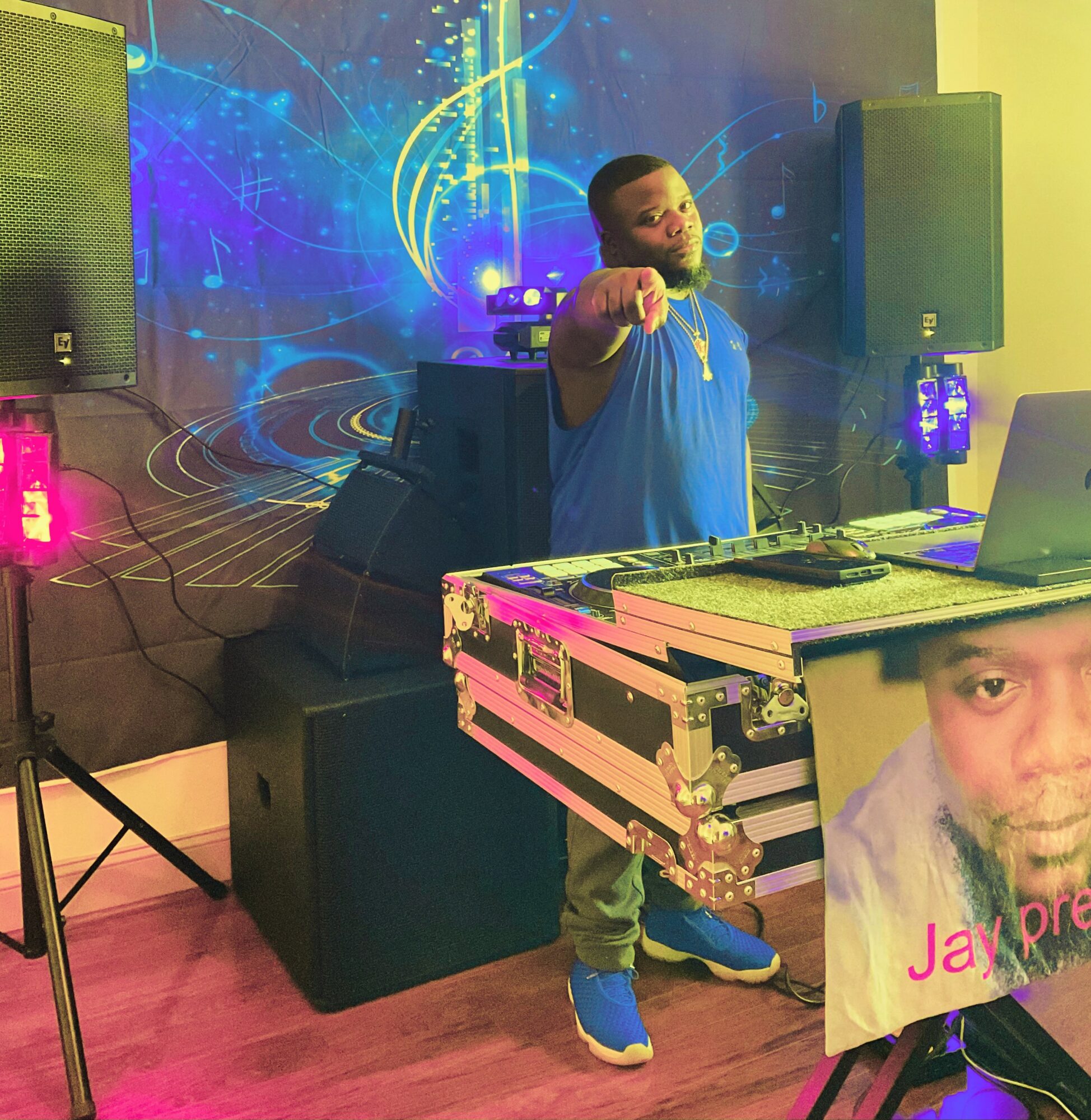 Person with a beard pointing at the camera, standing behind DJ equipment, with a colorful digital background, and a poster partially visible.