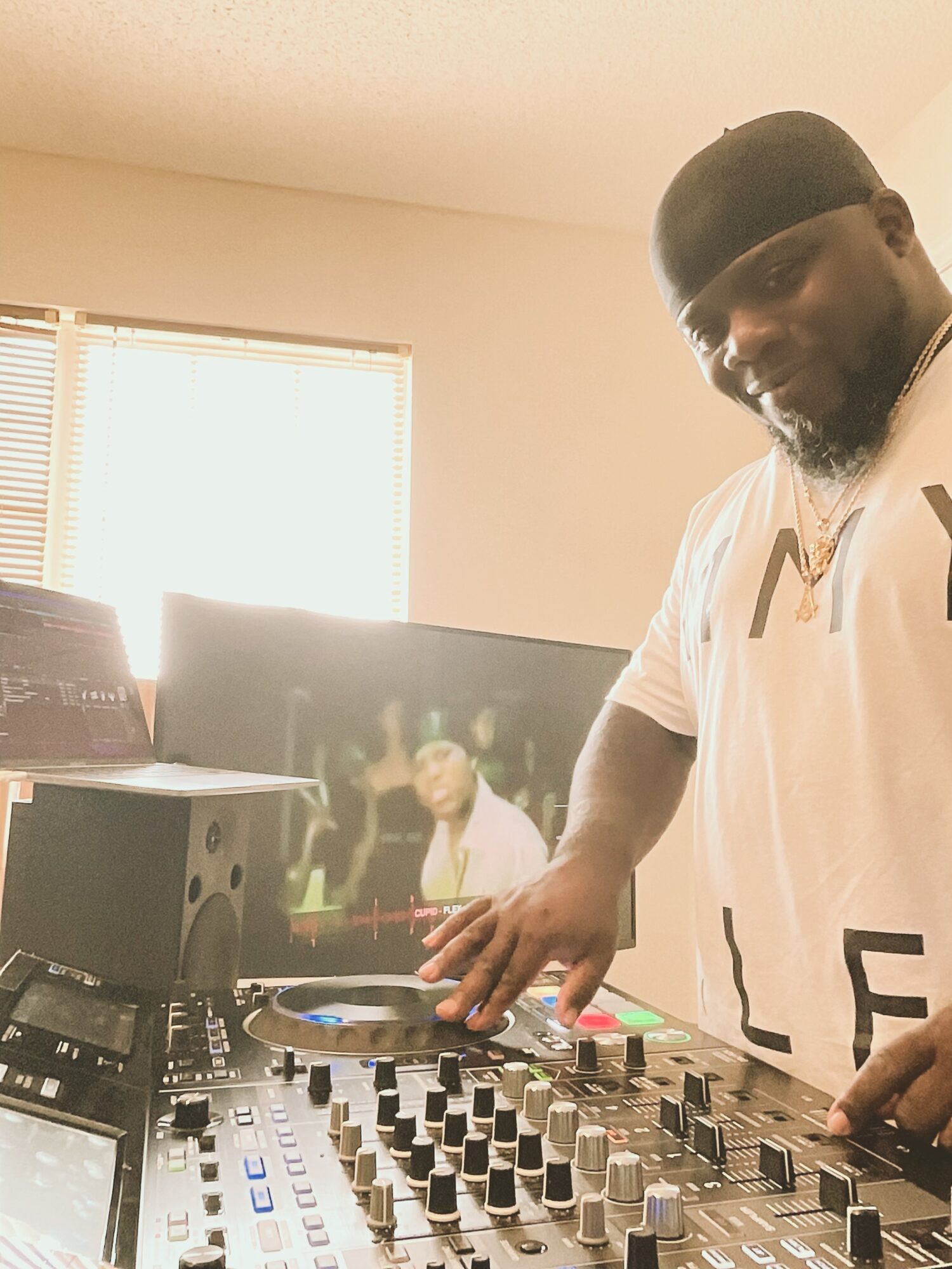 Man wearing a black cap and white shirt operating a DJ mixer in a room with a window and a television screen.